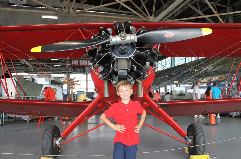 Andrew McMorris stands proudly before a red World War two airplane wearing a red shirt.