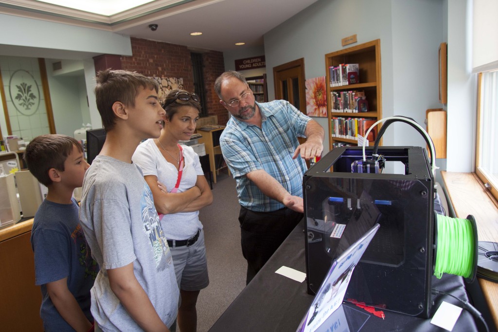 From left to right: Hunter Magnuson, 10, Ethan Magnuson, 12, and Kea Magnuson learn about the 3-D printer from library network administrator David Van Popering. (Credit: Chris Lisinski)